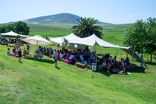 Outdoor event with people gathered under large tents on a grassy hillside with a mountain in the background.