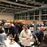 Winemakers presenting wines at the Nedbank CWG Auction Showcase in Cape Town with labeled banners overhead.