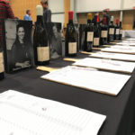 Table with wine bottles, portraits of winemakers, and bid sheets at the Nedbank Cape Winemakers Guild Auction Showcase.