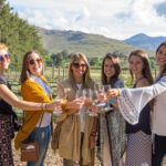 A group of six women smiling and holding wine glasses in a vineyard with mountains and greenery in the background.