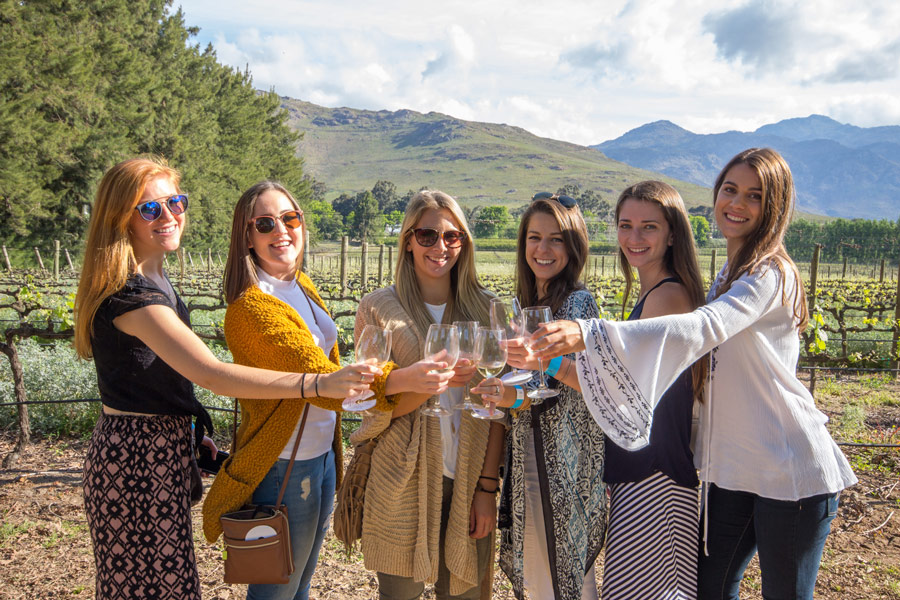 A group of six women smiling and holding wine glasses in a vineyard with mountains and greenery in the background.