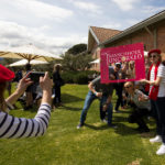 Group of friends posing with a Franschhoek Uncorked Festival frame, dressed in berets and scarves, while another person takes their picture in an outdoor setting with umbrellas and a brick building in the background.