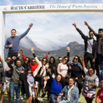 Large group of people posing cheerfully under a large white frame labeled 'Haute Cabrière: The Home of Pierre Jourdan' with a scenic mountain backdrop and cloudy sky.