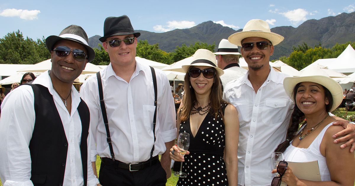 Group of five people enjoying an outdoor wine festival, smiling and dressed in stylish summer hats.