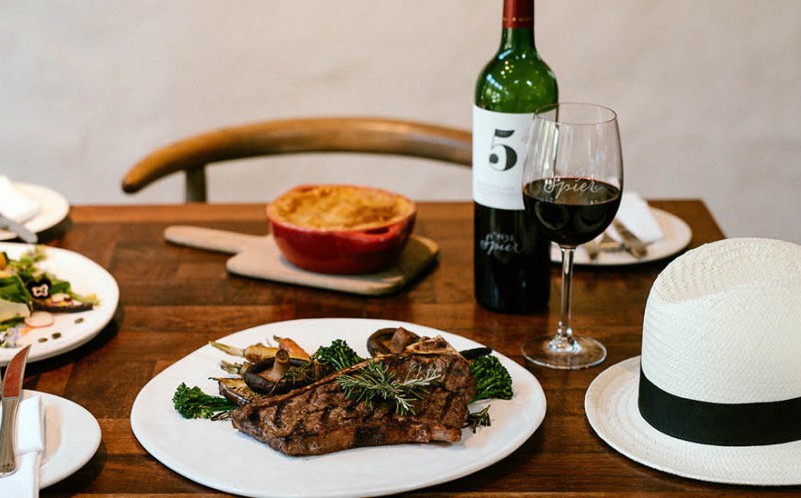 Elegant dining table setup with a steak dinner, a bottle of red wine, a filled wine glass, and a stylish white hat.