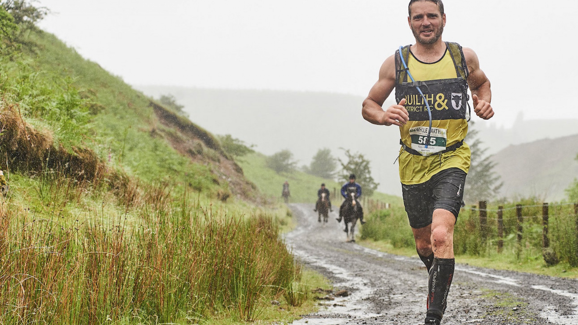 Trail runner navigating a muddy, rainy path in a green hilly landscape, with horseback riders in the distance.