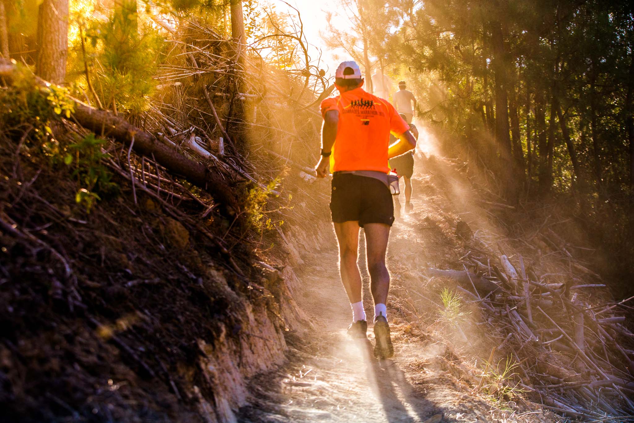 Trail runners in vibrant orange shirts racing through a sunlit forest trail, dust swirling around them.