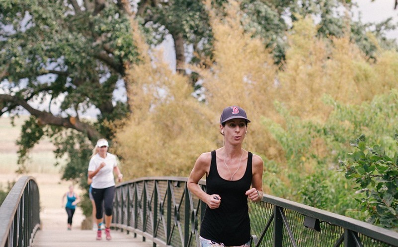 Three runners, with one prominently in the foreground, jogging across a bridge surrounded by lush greenery and autumn trees.